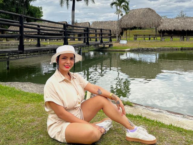 JessicaFiore with tan dress, white bucket hat, red lipstick, tattoos, and white sneakers, sitting by a pond, smiling, showing her ass, with green grass and palm trees.