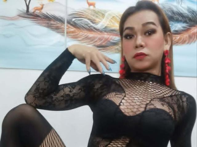 MarianeOhara with red earrings, black lace top, and fishnet sleeves, showing her tits through the lace, sitting with one leg up, blue nail polish, and a serious look.