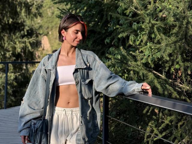 TameikaMacclellan with short brown hair, pink earrings, denim jacket, white tube top, and white pants, leaning on a black railing, standing beside green pine trees.