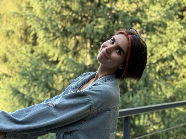 TameikaMacclellan with short black hair, red highlights, and a denim jacket, smiling slightly, eyes looking upwards, standing outdoors with a metal railing.