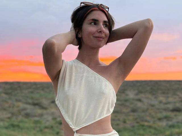 TameikaMacclellan with short dark hair, wearing a white one-shoulder top, arms raised, smiling, sunset sky, red headband, and sunglasses on top, in a grassy field.