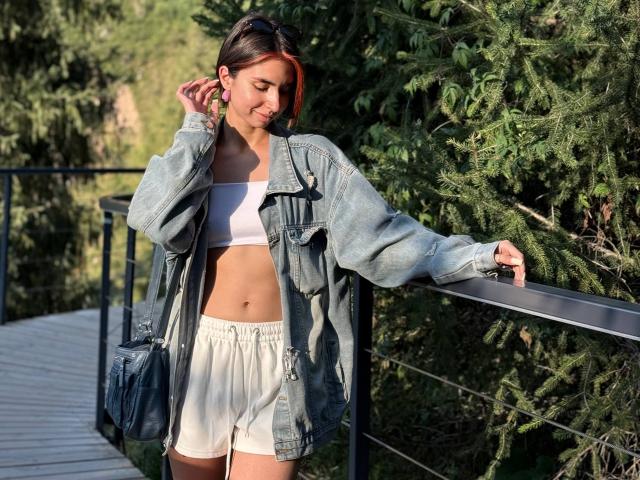 TameikaMacclellan, con el cabello teñido de rojo, vestida con una chaqueta vaquera, un top deportivo blanco y pantalones cortos color crema, apoyada en una barandilla, con la mano en el pelo, y un fondo de árboles verdes..