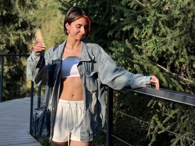 TameikaMacclellan stands on a wooden bridge, wearing a denim jacket over a white sports bra and white shorts, with pink earrings and a black bag. Her short brown hair.