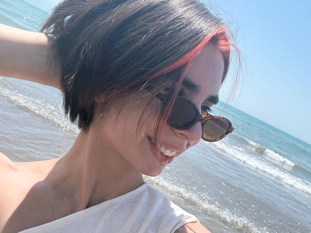 TameikaMacclellan with short black and red hair, wearing sunglasses, white top, smiling, showing teeth, by the ocean, hand in hair, clear blue sky, sunny, water in background.