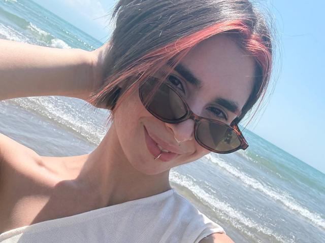 TameikaMacclellan has short brown and pink-dyed hair, wears tortoiseshell sunglasses, a white top, and a nose ring, smiling with a septum piercing at the beach.