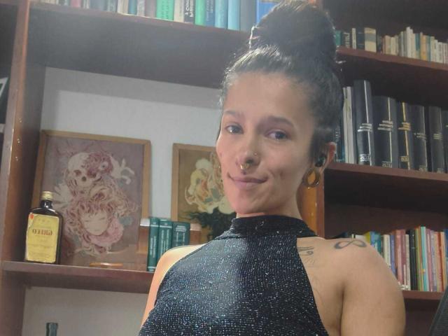 Amelie Blake, met donker haar in een knot, een neusring en gouden oorringen, draagt een zwarte haltertop en glimlacht, waarbij een tatoeage op haar rechter schouder zichtbaar is..