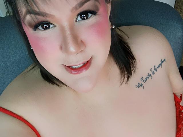 GiiannaMorrys with pink blush, brown eyes, and red lipstick, wearing red lingerie, showing a tattoo on her right shoulder, and a small nose ring. Her tits are partially visible.