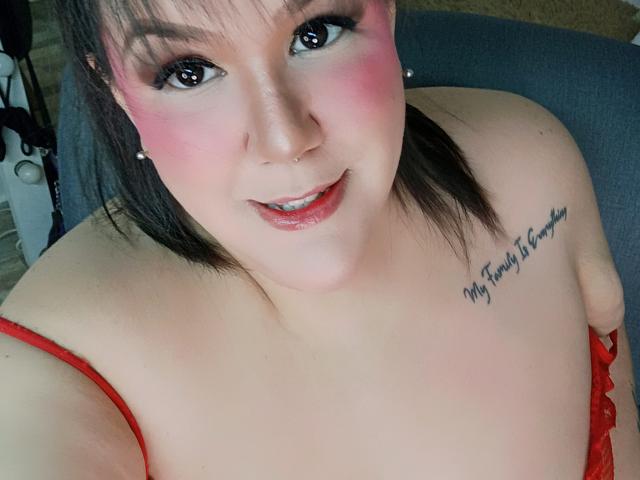 GiiannaMorrys has pink blush, black eyeliner, and a nose piercing. She's smiling with red lipstick, wearing red lingerie, showing her tits, and has a tattoo on her right chest.