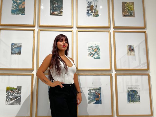 AlyssaGreen with long brown hair, white tank top, and black pants stands in front of a wall of framed pictures. She's looking at the camera, hand in pocket.