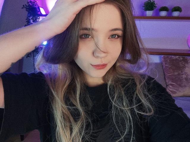 CarylPetru with long brown hair, light skin, and green eyes, wearing a black shirt, touching her head, looking at the camera with a slight smile.