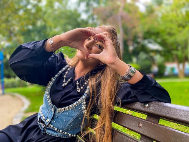 EveBliss with long brown hair, wearing a black shirt and studded denim vest, making a heart shape with her hands, sitting on a wooden bench, looking cute.