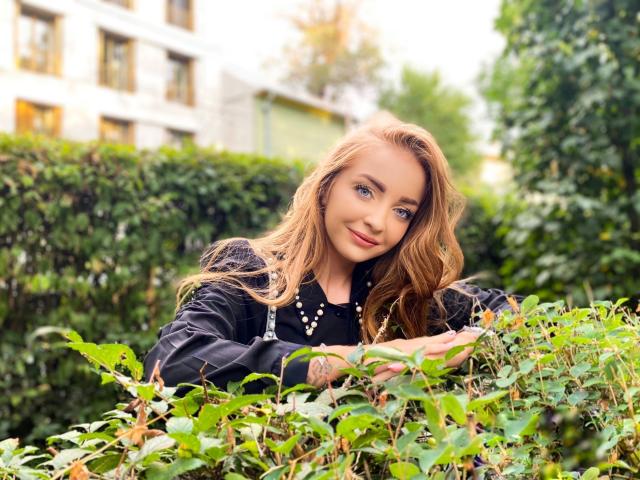 EveBliss with long blonde hair, blue eyes, black top, and pearl necklace, smiling while leaning on green bushes, hands resting on leaves, outdoor setting, daytime.