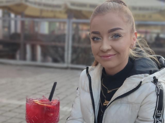 EveBliss with blonde ponytail, blue eyes, and slight smile, wearing white jacket and black shirt, holding red drink with straw, sitting outside.