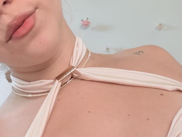 SugarrX's face, showing pink lips, a small heart tattoo on the right shoulder, and a pink halter top with a gold clasp, exposing her collarbone and neck.