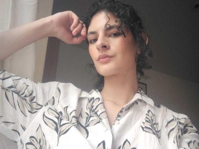 RoxyGraff has curly black hair, light skin, and is wearing a white floral shirt. She's looking down with a slight smile, touching her head with her right hand.