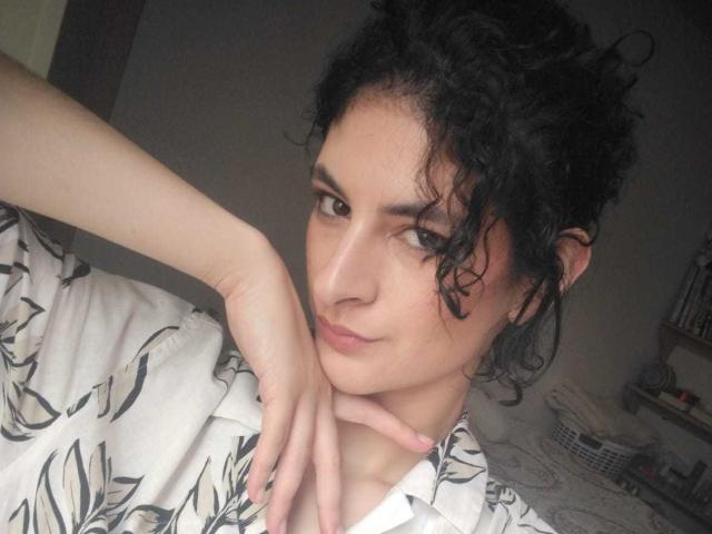 RoxyGraff with curly black hair, light skin, and subtle makeup, wearing a white floral shirt, resting her chin on her hand, and giving a slight, confident smile.