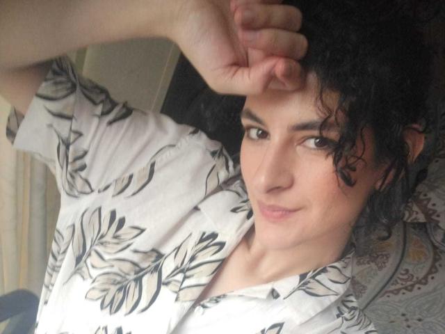 RoxyGraff with curly black hair, wearing a white floral shirt, raising her arm to cover her forehead, looking at the camera with a slight smile.