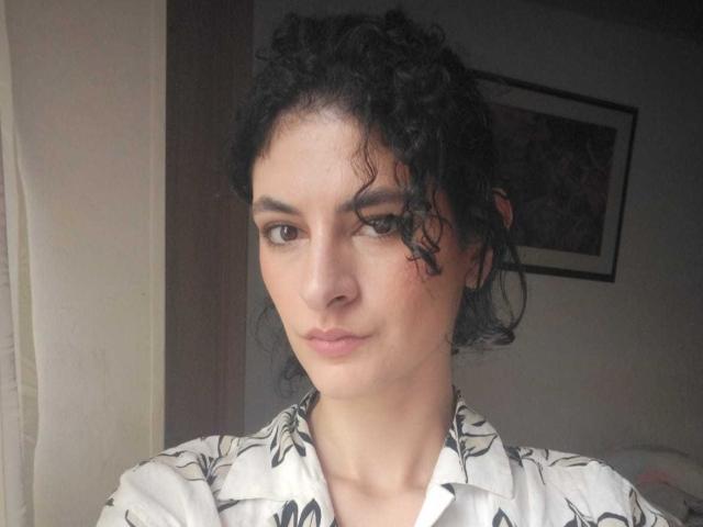 RoxyGraff has curly black hair, light skin, and brown eyes, wearing a white floral shirt. She's looking slightly to the side with a neutral expression.