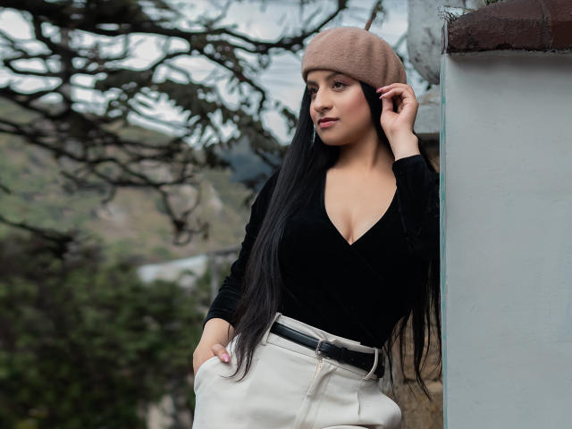 ElyThemsom, con lunghi capelli neri, indossa un berretto beige, una maglia nera a V e pantaloni bianchi, si tocca l'orecchio, è in piedi vicino a un muro bianco, mostrando piccoli seni..