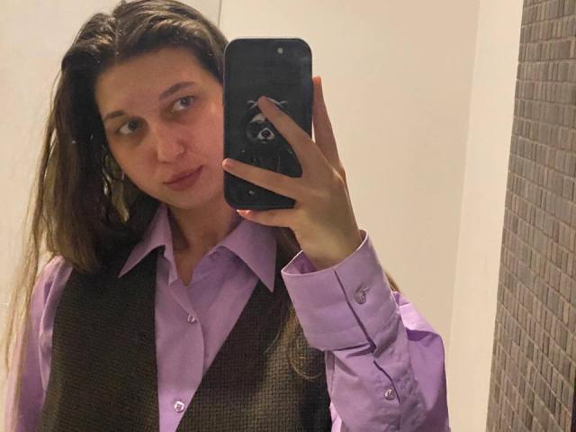 Sanella with long brown hair, wearing a purple shirt and brown vest, taking a mirror selfie with a black phone, showing a neutral expression.
