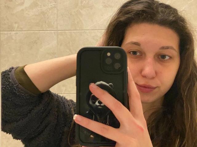 Sanella with long brown hair, wearing a fuzzy dark blue shirt, holding a black phone with a black pop socket, taking a selfie with a pouty expression.