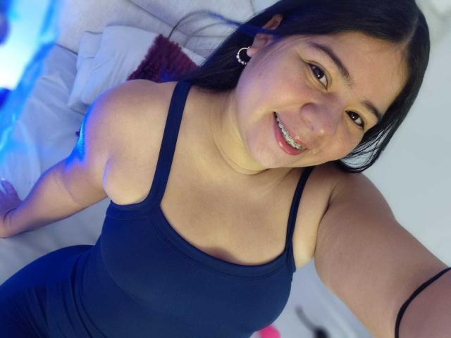 ElizaWiliamst with dark hair, smiling, wearing a black tank top, showing her tits, small hoop earrings, and black bracelet, sitting on a bed with blue lighting.