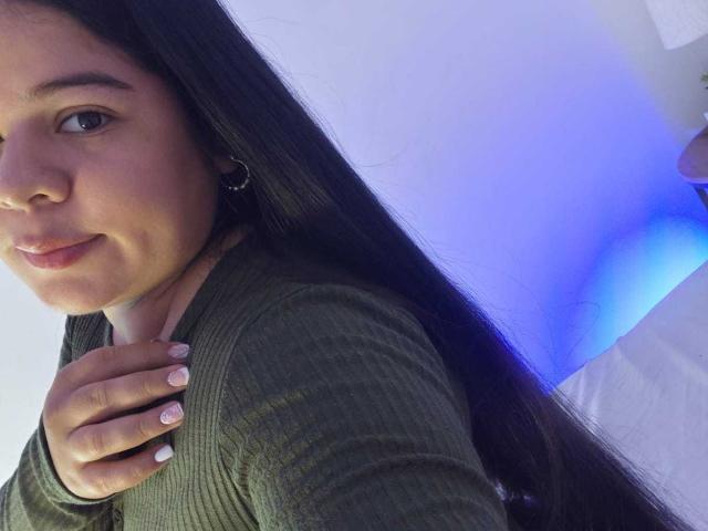 ElizaWiliamst with long black hair, green ribbed shirt, and white nails, hand on chest, smiling slightly, blue light on right. Her ass is not visible.