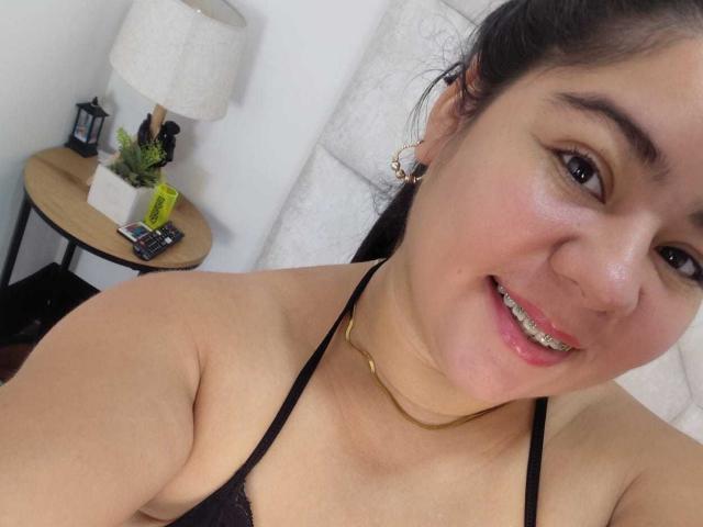 ElizaWiliamst with a smile, black halter top, gold chain necklace, and earrings, showing her tits, dark hair, and shiny lips, standing near a small wooden table.