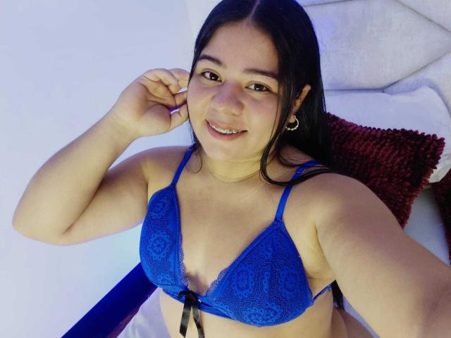 ElizaWiliamst with dark hair, smiling, wearing a blue lace bra, showing her tits, hand near ear, small hoop earrings, red pillow in background, white towel.