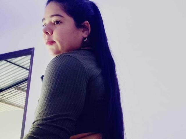 ElizaWiliamst con lunghi capelli neri, una maglia a maniche lunghe verde a coste e orecchini, che si guarda indietro, mostrando la schiena e i glutei, in piedi vicino a uno specchio..