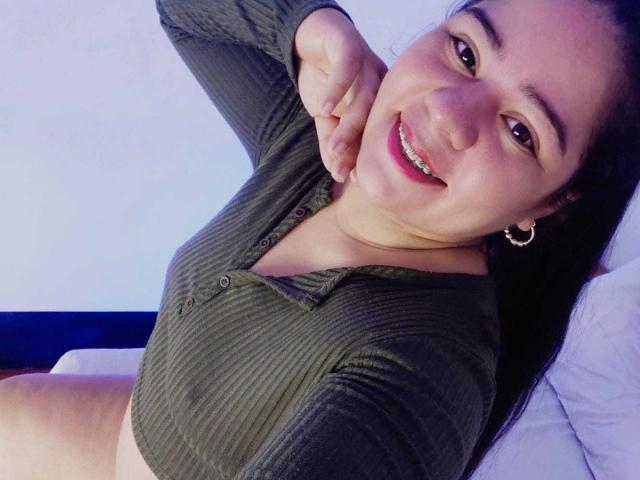 ElizaWiliamst con una camicia a coste verde scuro, sorridente, che mostra i denti, con una mano al collo, piccoli orecchini a cerchio, sdraiata su un cuscino bianco, con il seno visibile, capelli lunghi neri, pelle chiara, labbra rosa..