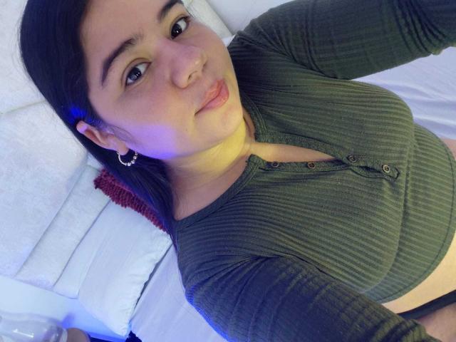 ElizaWiliamst with dark hair, green ribbed long-sleeve shirt, small nose ring, and hoop earrings, lying on bed, showing her tits and a bit of her ass.