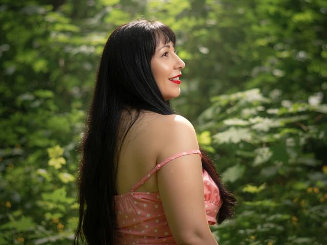 LiluGrants with long black hair, red lipstick, and pink polka dot top, smiling over her shoulder, showing her ass, in a sunlit forest.