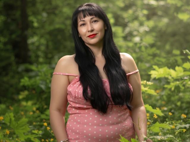 LiluGrants has long black hair, red lipstick, and a pink polka dot dress with off-shoulder straps, standing in a green forest with yellow flowers, smiling softly.