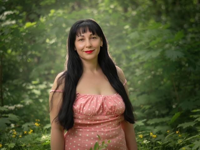 LiluGrants with long black hair, red lipstick, and pink polka-dot dress, showing her tits, smiling in a green forest. She's cute and has a curvy, sexy body.