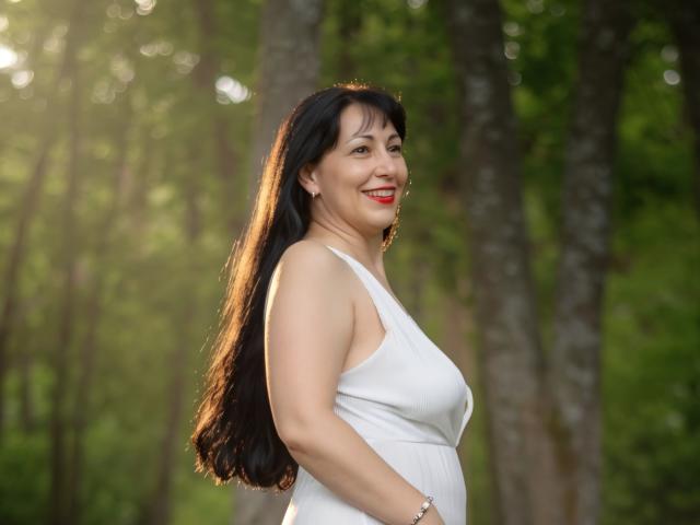 LiluGrants with long black hair, smiling, wearing a white sleeveless dress, showing her tits, standing in a sunlit forest. She's got red lipstick and a silver bracelet.