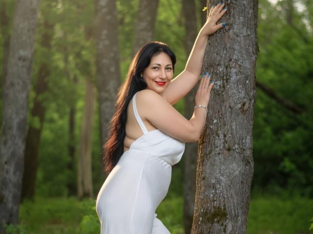 LiluGrants with long black hair, red lipstick, and a white dress, leans against a tree, showing her ass, smiling, with blue nails, in a forest.