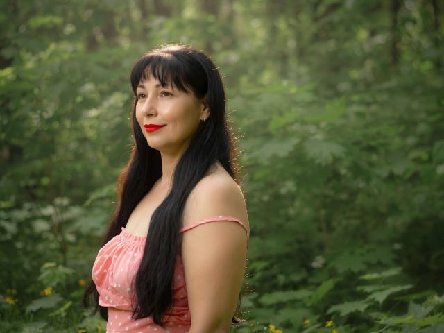 LiluGrants with long black hair, red lipstick, and a pink off-shoulder dress, standing in a forest, showing her cute ass and small tits.