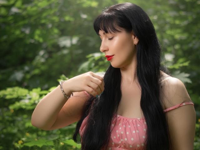 LiluGrants with long black hair, red lipstick, and a pink polka-dot top, touching her hair, showing her ass and tits, in a forest with green foliage.