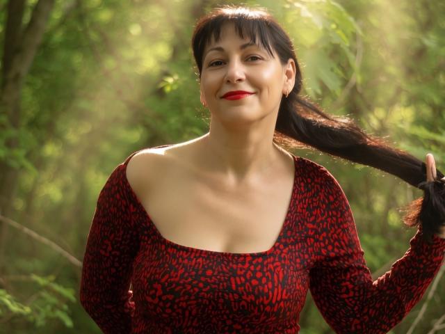 LiluGrants with red leopard print dress, holding black hair, smiling, red lipstick, medium tits, light skin, green forest background, sunlight, holding hair in right hand.
