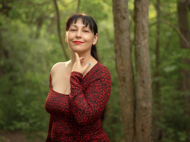 LiluGrants with black hair and red patterned dress, smiling, hand on neck, showing bare shoulder, blue nail, forest background. Boobs partially visible. Red lipstick. Casual, confident.