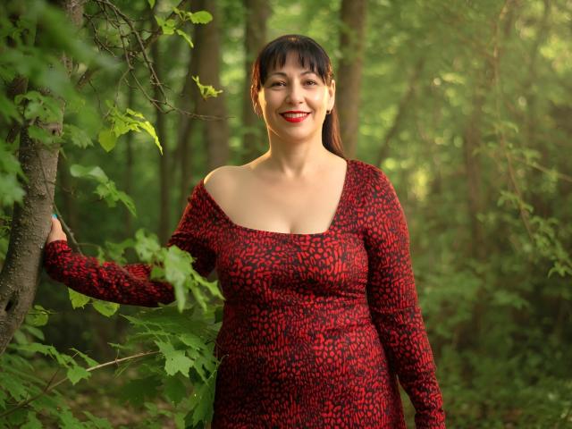 LiluGrants stands in a forest, wearing a red and black patterned dress, with her hand on a tree, smiling, showing red lipstick, and having medium-sized boobs.