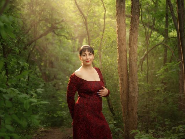 LiluGrants standing in a forest, wearing a red, long-sleeved dress with a floral pattern, hand on her chest, smiling, surrounded by tall trees and green foliage.