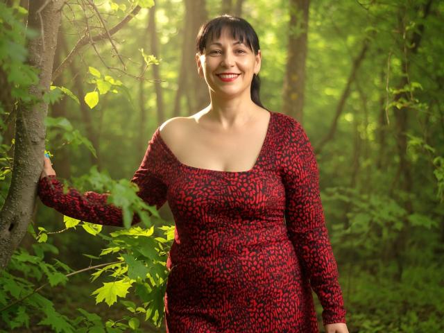LiluGrants stands in a forest, wearing a red and black patterned dress, with short black hair, smiling, and holding a tree with green leaves and sunlight.