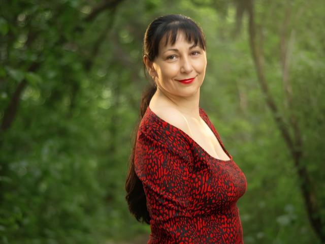 LiluGrants has red and black patterned top, medium tits, dark hair, red lipstick, smiling, standing in forest, green background, soft light, looking at camera.
