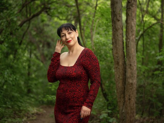 LiluGrants with black hair, red lipstick, and red leopard print dress, standing in a forest, smiling, touching her ear, showing her tits, with a hand on her hip.