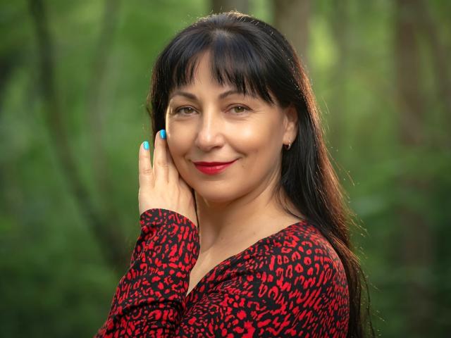LiluGrants mit langen, schwarzen Haaren, einem roten Top mit Leopardenmuster, blauer Nagellack, rotem Lippenstift und einem leichten Lächeln, die ihre Wange in einem Wald berührt. Ihre Brüste sind klein..
