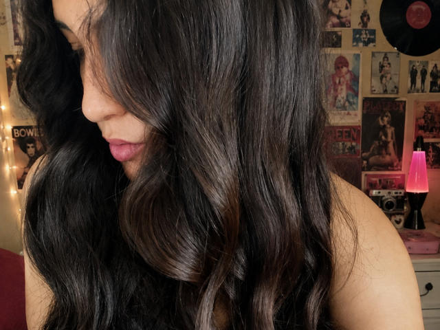 MargaritaDudole with long, wavy, black hair, shiny and voluminous, covering her face and shoulder. Pink lipstick, soft brown skin, and a pink lamp in the background.