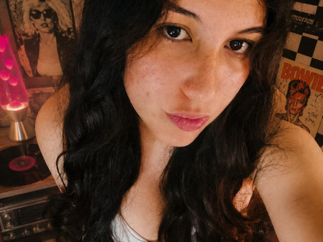 MargaritaDudole with wavy black hair, freckles, and pink lips, looking into the camera, wearing a white tank top, showing her tits, with a pink dildo in the background.