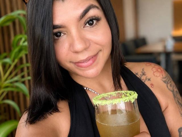 ValeryPierce with dark hair, brown eyes, and tattoos, smiling while holding a green-sugared drink in a glass. Wearing a black halter top, pearl necklace.